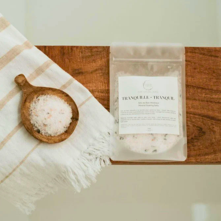 Un sac de sel de bain avec l'inscription tranquille - tranquil  sur une planche de bois.  À sa gauche un vois un petit récipient de sel de bain déposé sur une serviette.