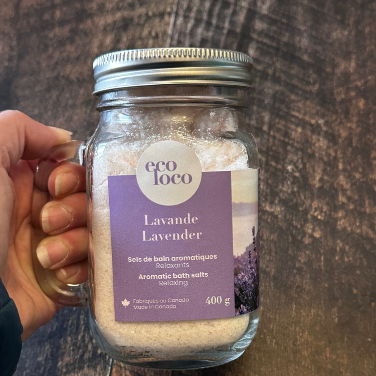 Un sel de bain à la lavande dans un pot Mason avec poignée tenu dans une main au dessus d'une planche de bois