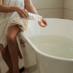 Une femme revêtu d'une serviette mettant du sel de bain dans une baignoire blanche remplie d'eau. 