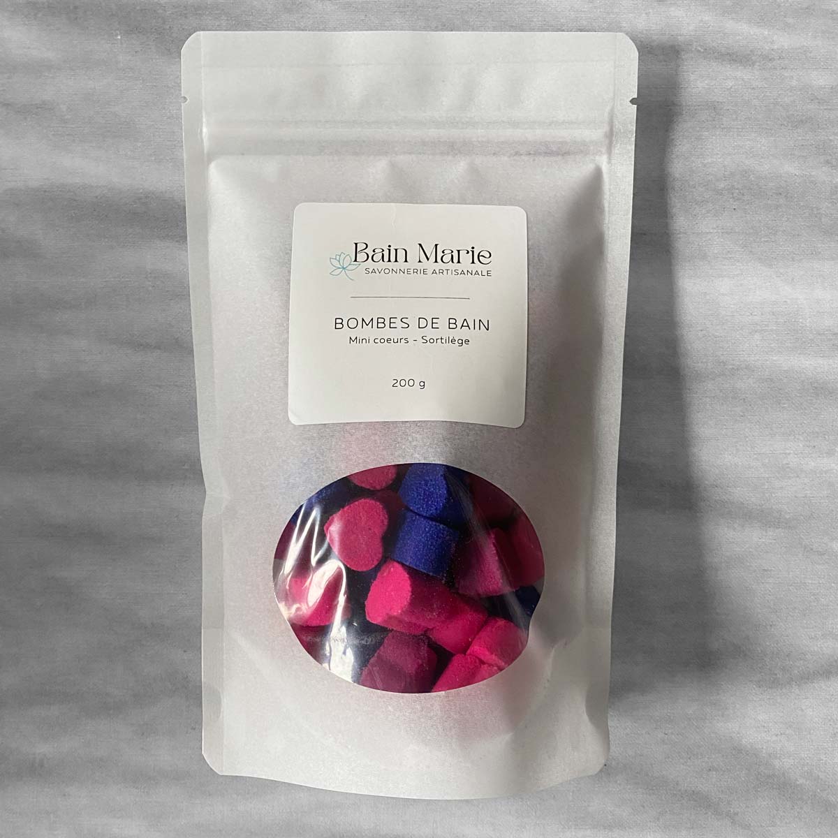 sac blanc avec une fenêtre qui laisse paraitre des petites coeurs roses, magentas et mauves sur un fond blanc effet de marbre gris. Étiquette blanche avec les inscriptions Bain Marie Savonnerie artisanale Bombes de bain mini coeurs sortilège 200g