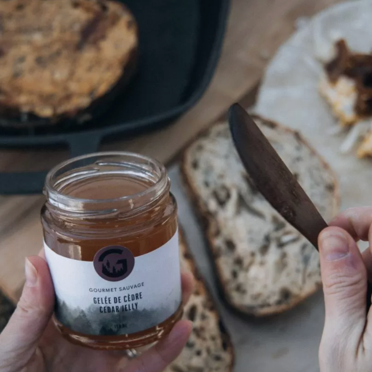 Pot de gelée de cèdre de Gourmet sauvage tenue dans une main de femme au-dessus d'un plateau de pain.  Vendu par Eco Loco