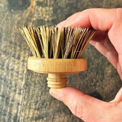 Tête de remplacement pour un pommeau ou une brosse  vaisselle en bois avec des poils durs tenu dans une main au-dessus d'une planche de bois. 