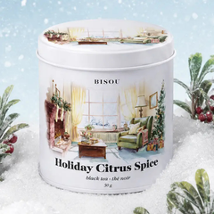 Une boite ronde et blanche avec un dessin d'un salon chaleureux comprenant une chaise verte et un foyer et des inscription Bisou Holiday Citrus Spice entouré de neige et de plantes. 