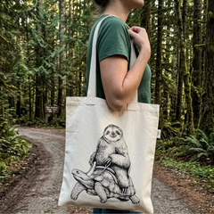 Une femme dans un chemin en forêt avec un chandail vert portant sur l'épaule un sac fourre-tout ayant comme motif un paresseux sur le dos d'une tortue. 