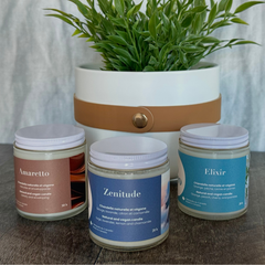 Trio de chandelle avec des étiquettes de différentes couleurs sur lesquelles on peut lire:  Amaretto, Zenitude et Elixir en avant d'un pot blanc contenant une plante verte. 