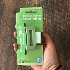 Petite pince à cheveux de couleur jade avec un logo de matériau recyclé et son emballage cartonné vert qui dit:  C'était un pot de pilules, créée pour tenir pas pour jeter, Unique plastique et le logo fait au Québec au-dessus d'une blanche en bois.  