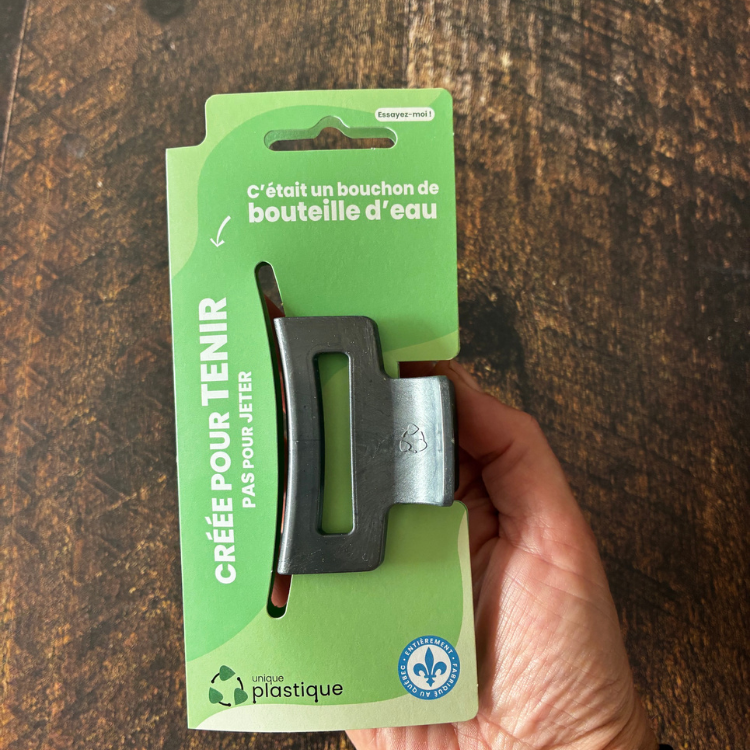 Petite pince à cheveux de couleur charbon avec un logo de matériau recyclé et son emballage cartonné vert qui dit:  C'était un bouchon de bouteille d'eau, créée pour tenir pas pour jeter, Unique plastique et le logo fait au Québec au-dessus d'une blanche en bois. 