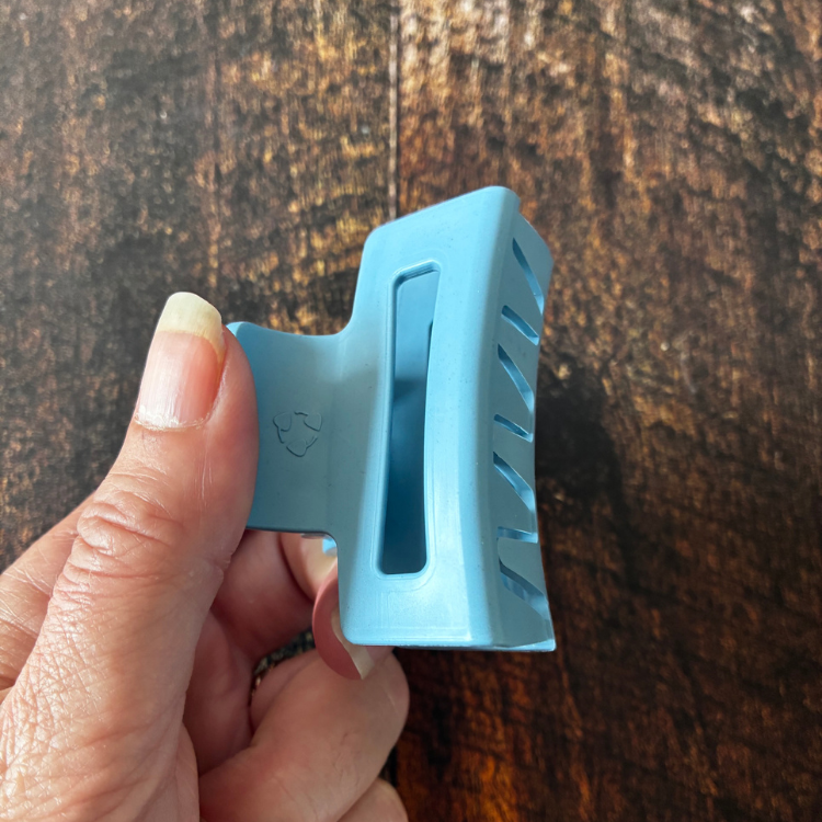 Petite pince à cheveux bleue avec le logo de matériau recyclé tenu dans une main féminine au-dessus d'une planche de bois. 
