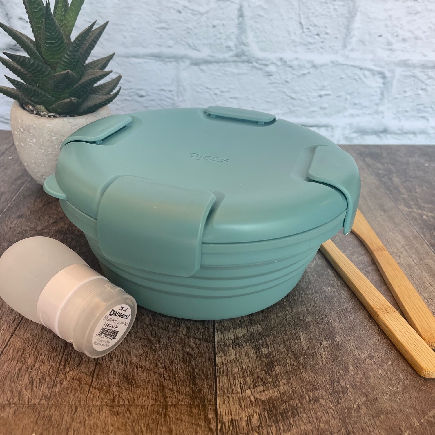 bol en silicone rétractable de couleur eucalyptus avec un couvercle comprenant 4 clips sur le couvercle déposé sur une blanche de bois avec des ustensiles en bois, une petite plante et un arrière plan de brique blanche. 