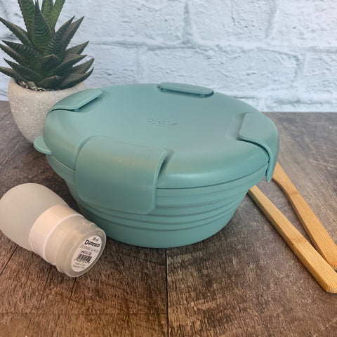 bol en silicone rétractable de couleur eucalyptus avec un couvercle comprenant 4 clips sur le couvercle déposé sur une blanche de bois avec des ustensiles en bois, une petite plante et un arrière plan de brique blanche. 