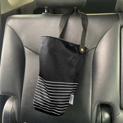 Corbeille poubelle en tissus noir avec des lignes blanches et une ganse noire avec une étiquette blanche Eco Loco qui est accrochée à la tête d'un siège de voiture adossée sur des bancs de cuir noir.