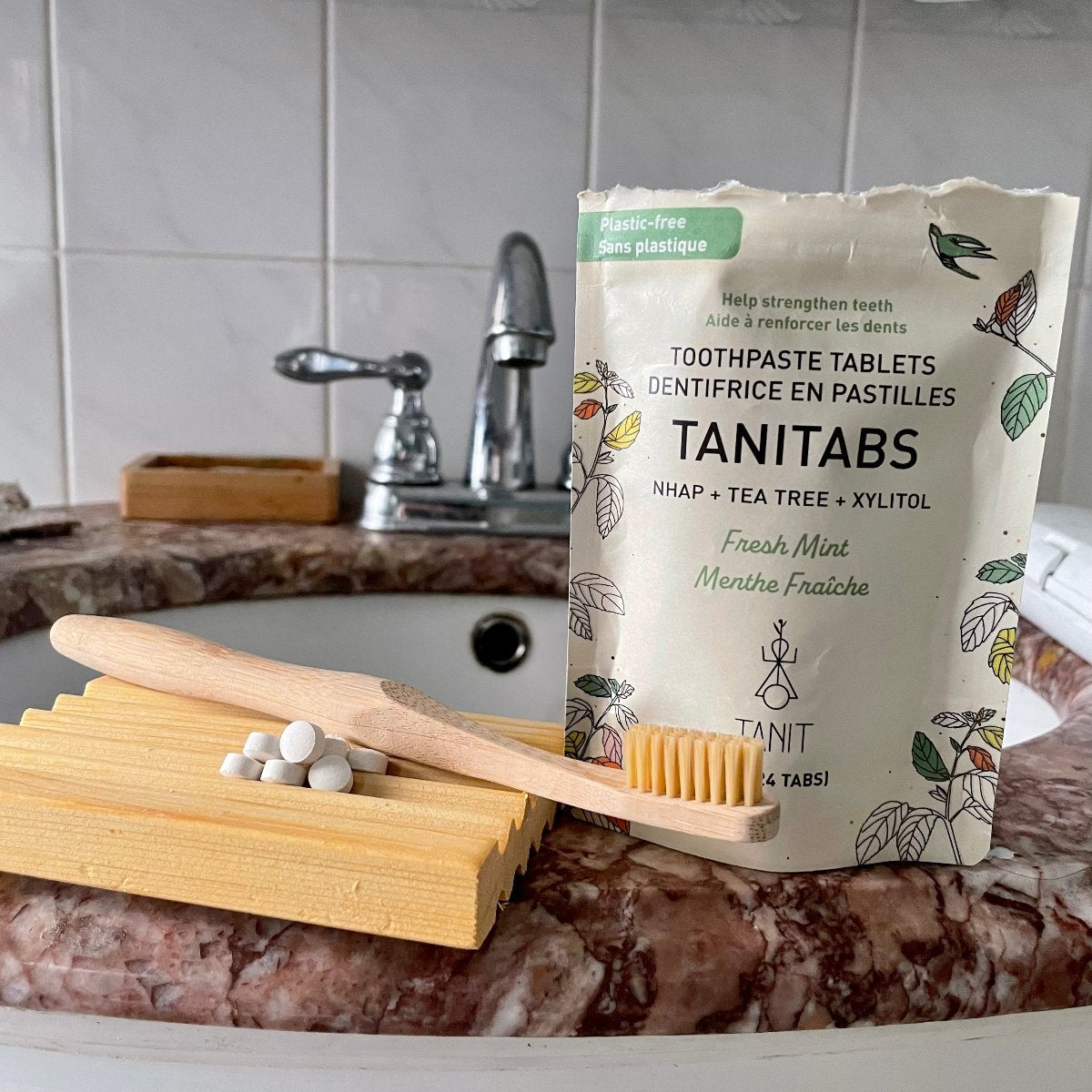 Un sachet de dentifrice en pastilles sur lequel il est écrit Tanitab menthe fraiche Tanit aide à renforcer la dent déposé sur un lavabo avec un porte savon et une brosse à dent en bambou. 